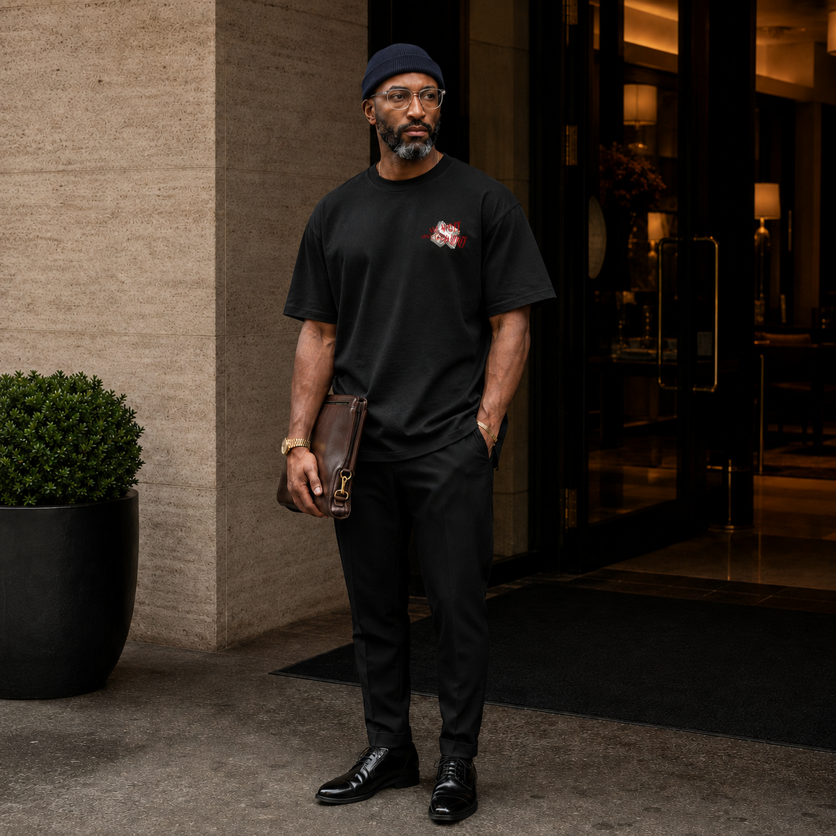 A man wearing a black crew-neck t-shirt with a small graphic on the left chest, black trousers, and black leather shoes stands in front of a hotel entrance holding a brown leather clutch.