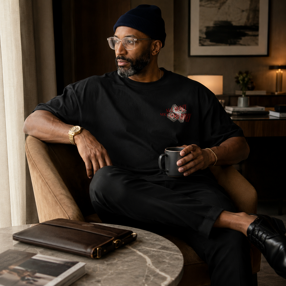 A man wearing a black beanie, glasses, and a black graphic t-shirt sits in a brown armchair holding a dark gray mug. The room has warm, ambient lighting, a marble side table with a brown leather portfolio case, and framed wall art in the background.