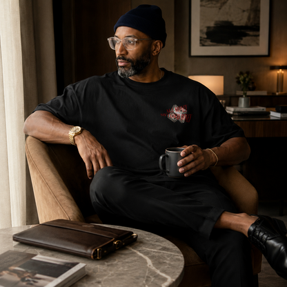 A man wearing a black beanie, glasses, and a black graphic t-shirt sits in a brown armchair holding a dark gray mug. The room has warm, ambient lighting, a marble side table with a brown leather portfolio case, and framed wall art in the background.