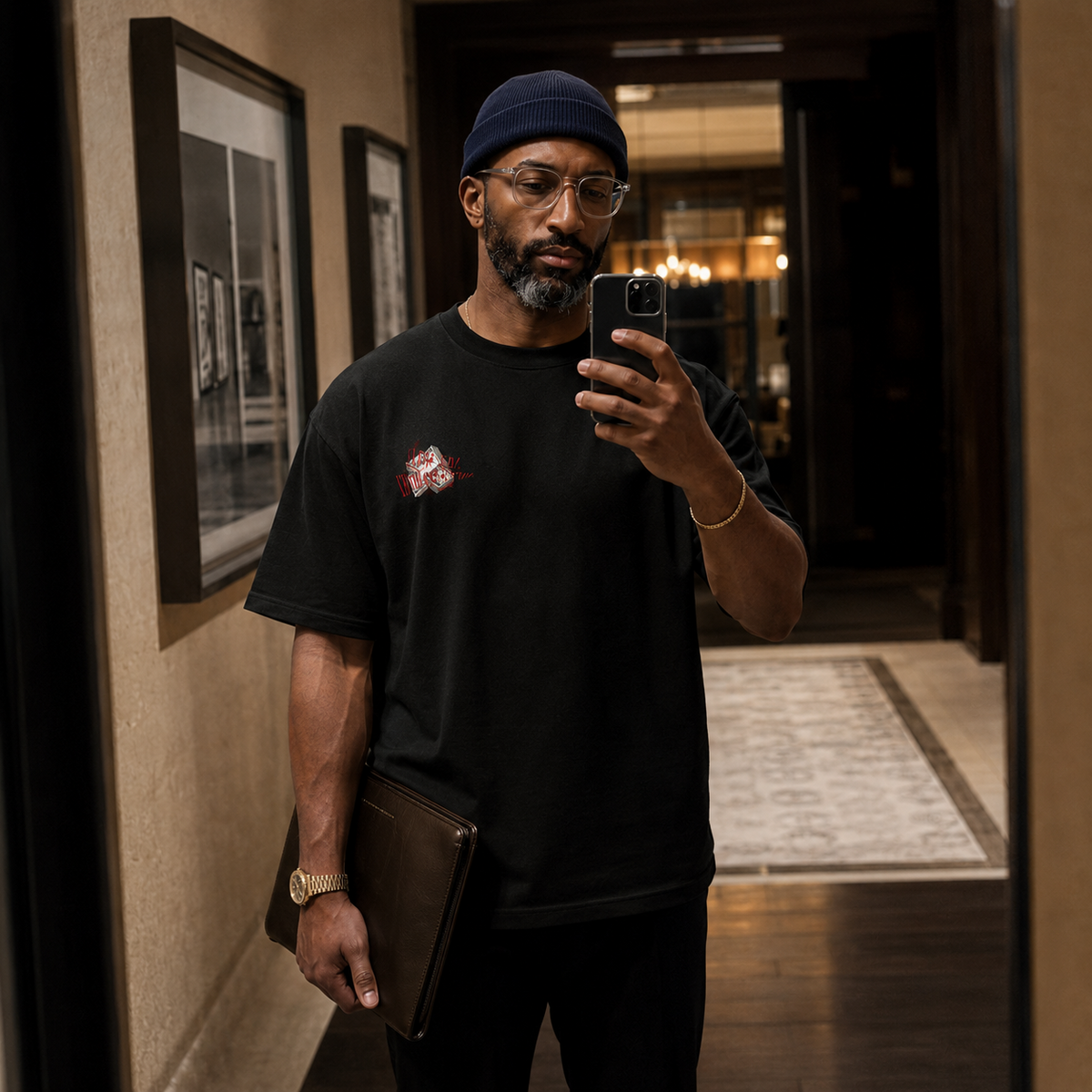 A man takes a mirror selfie in a hallway, wearing a black crew-neck t-shirt with a small red and white graphic on the left chest, a navy blue beanie, and holding a laptop.