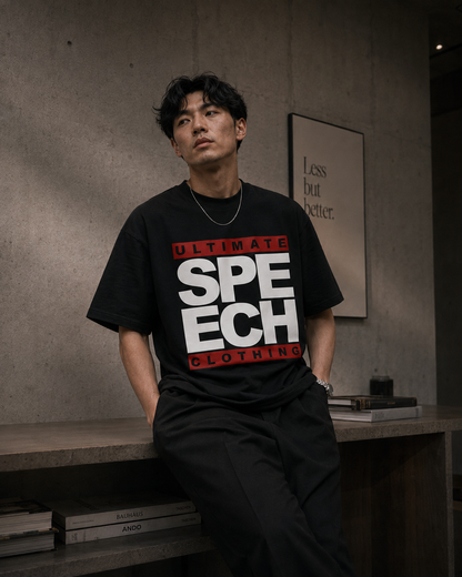 A man wearing a black graphic t-shirt with 'ULTIMATE SPEECH CLOTHING' printed in bold white and red letters, sitting on a wooden bench against a concrete wall.