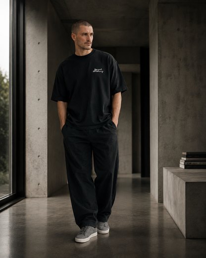 A man wearing a black crewneck t-shirt with a small white script logo on the chest, paired with black sweatpants and gray sneakers, standing in a modern concrete hallway.