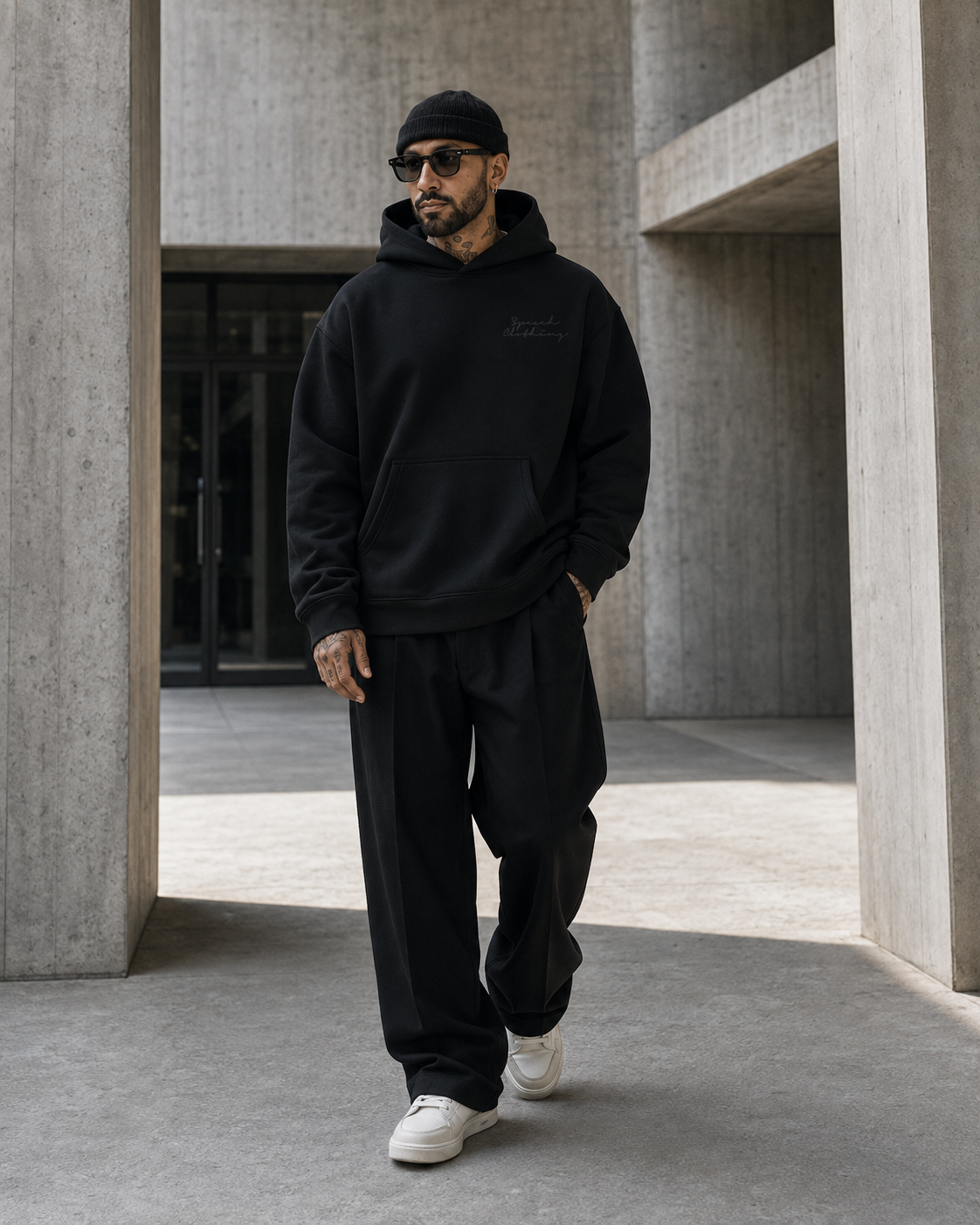 A man wearing a black hoodie with a small embroidered logo on the chest, black sweatpants, a black beanie, and white sneakers, walking through a modern concrete corridor.