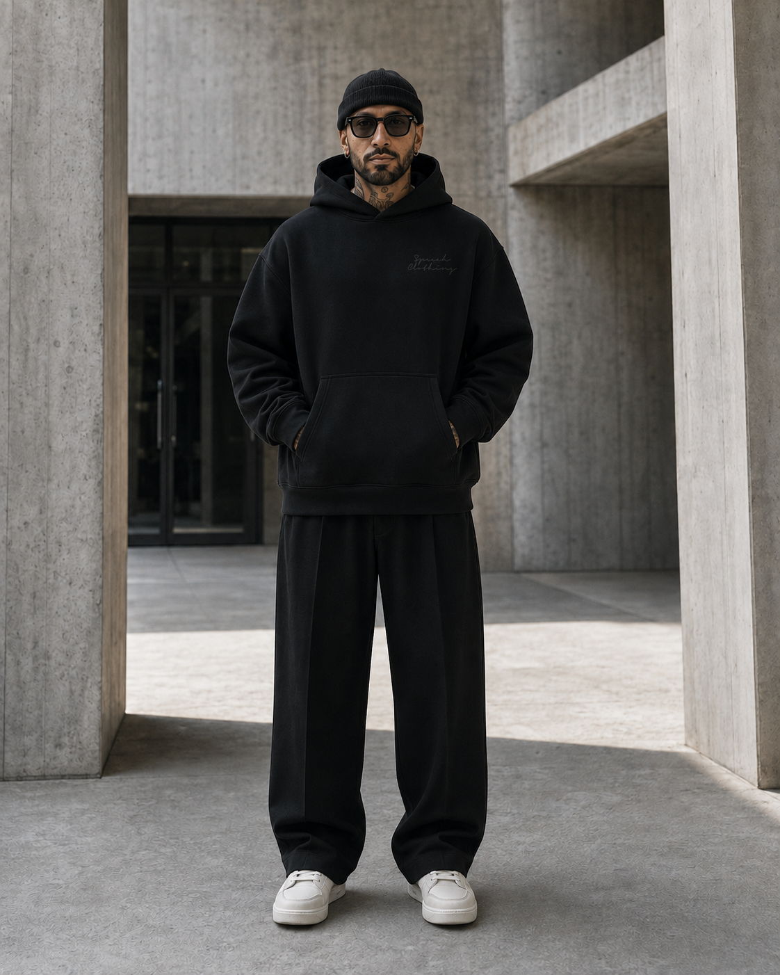 A man wearing a black pullover hoodie, black sweatpants, a black beanie, and white sneakers stands in a concrete architectural corridor.