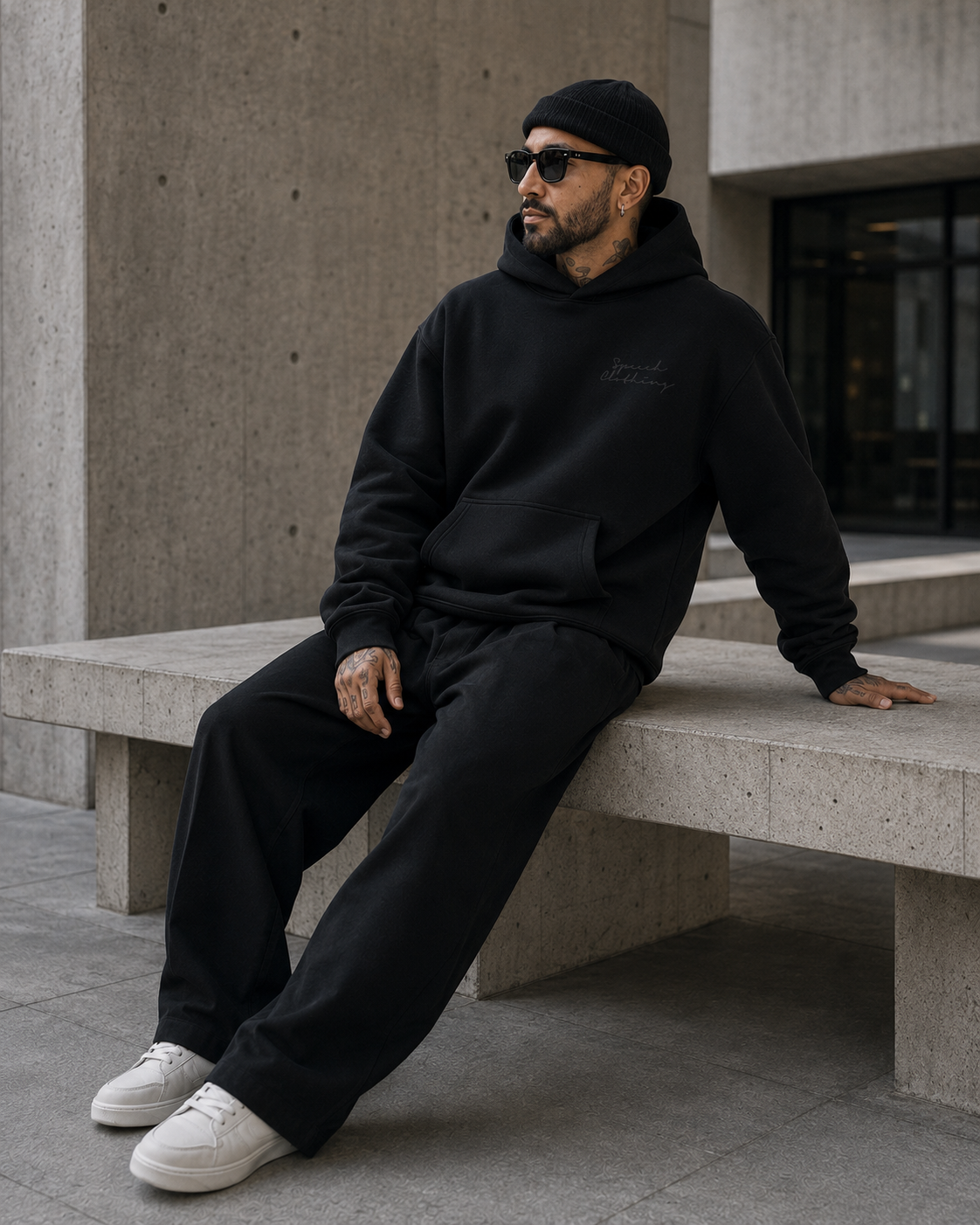 A man wearing a black hoodie, black sweatpants, a black beanie, and white sneakers sits on a concrete bench in an outdoor urban setting.