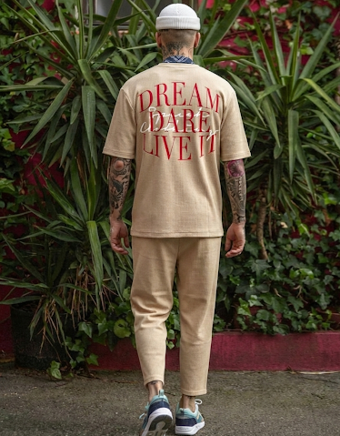 Person wearing a beige t-shirt with 'Dream Dare Live It' text on the back, standing in front of green plants.