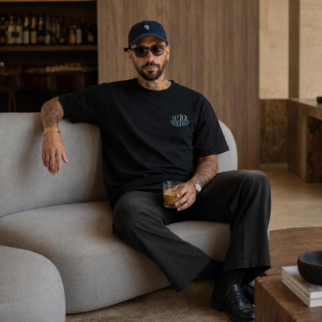 A man wearing a black t-shirt with a teal graphic and a matching black cap sits on a light gray sofa in a modern interior setting.