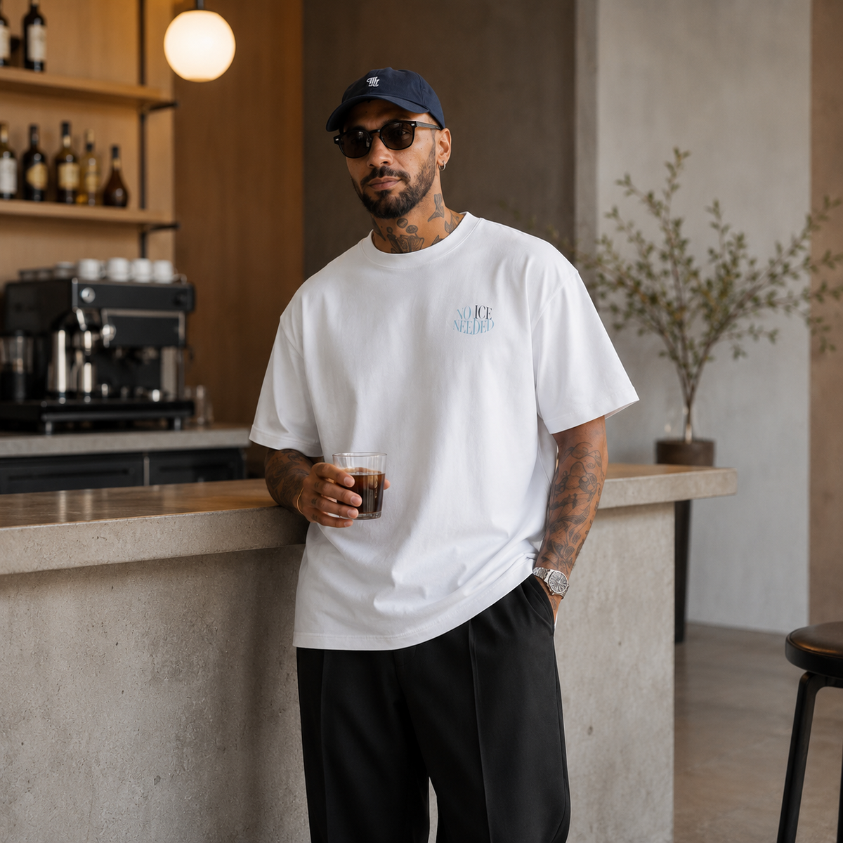 A man wearing a white crewneck t-shirt with a small light blue graphic on the left chest, black pants, and a navy blue baseball cap, standing in a modern cafe setting.