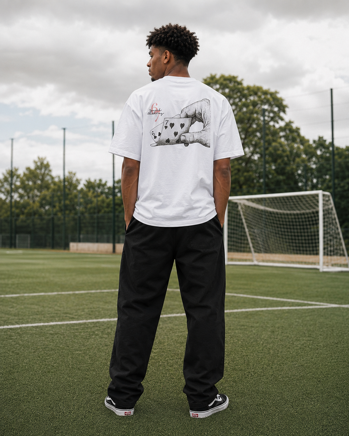 A man stands on an outdoor soccer field wearing a white t-shirt with a large graphic print on the back, black pants, and black sneakers, viewed from behind.
