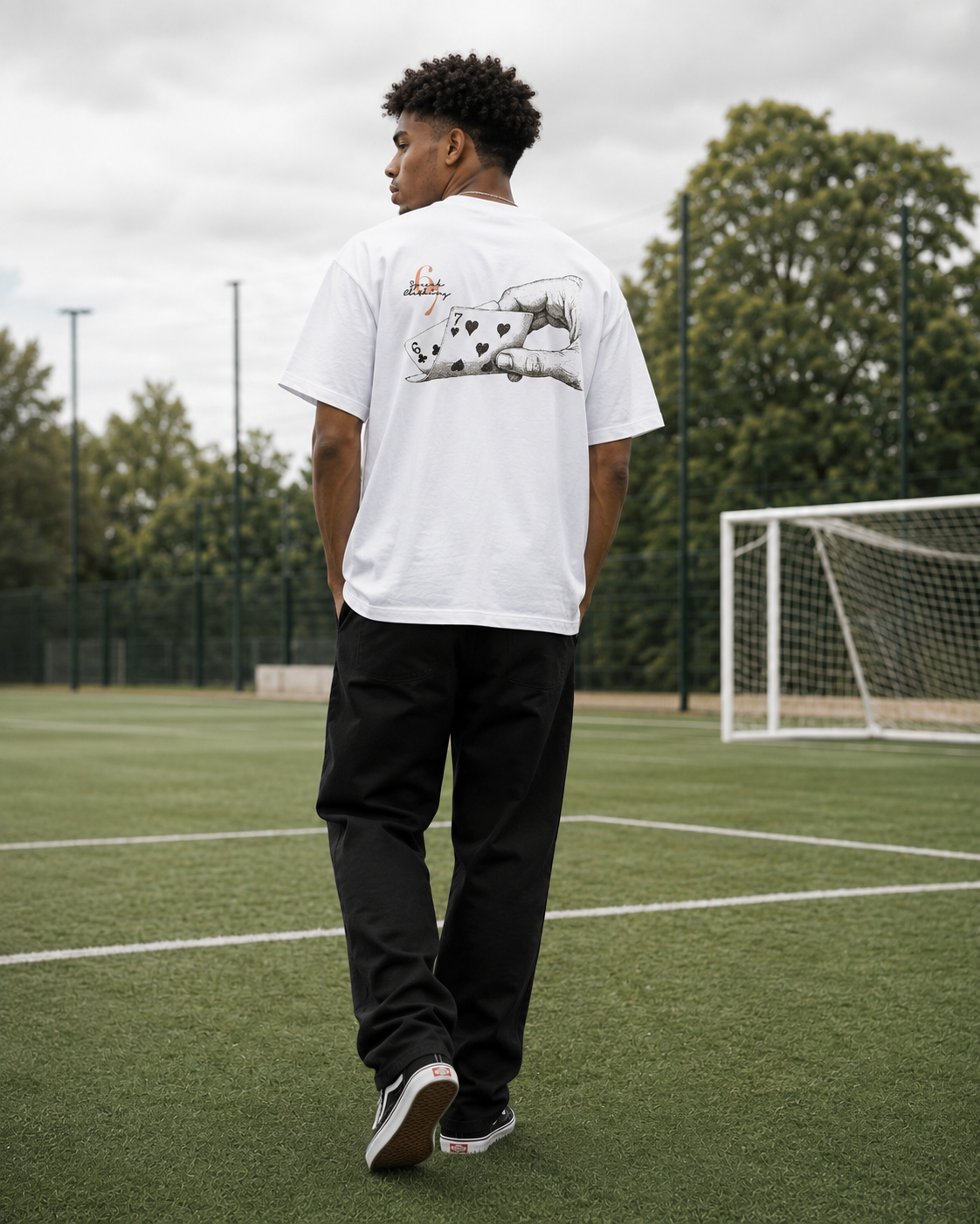 A man standing on an outdoor soccer field, viewed from behind, wearing a white t-shirt with a large graphic print on the back, black sweatpants, and sneakers.