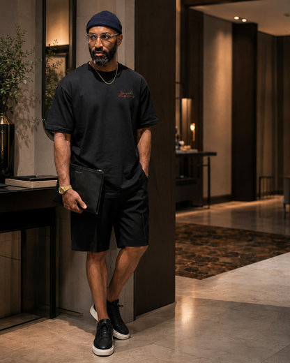 A man wearing a black crewneck t-shirt, black shorts, and black sneakers with white soles stands in a modern hotel lobby holding a black clutch.
