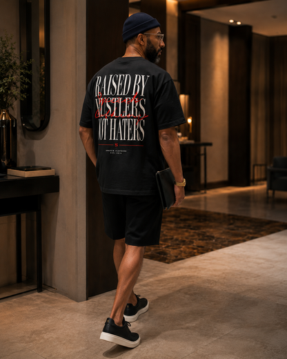 A man wearing a black graphic t-shirt, black shorts, and a navy blue beanie walks through a modern indoor hallway. The back of the t-shirt features a large graphic print with the text 'RAISED BY WRESTLERS OT HATERS' in white and red.