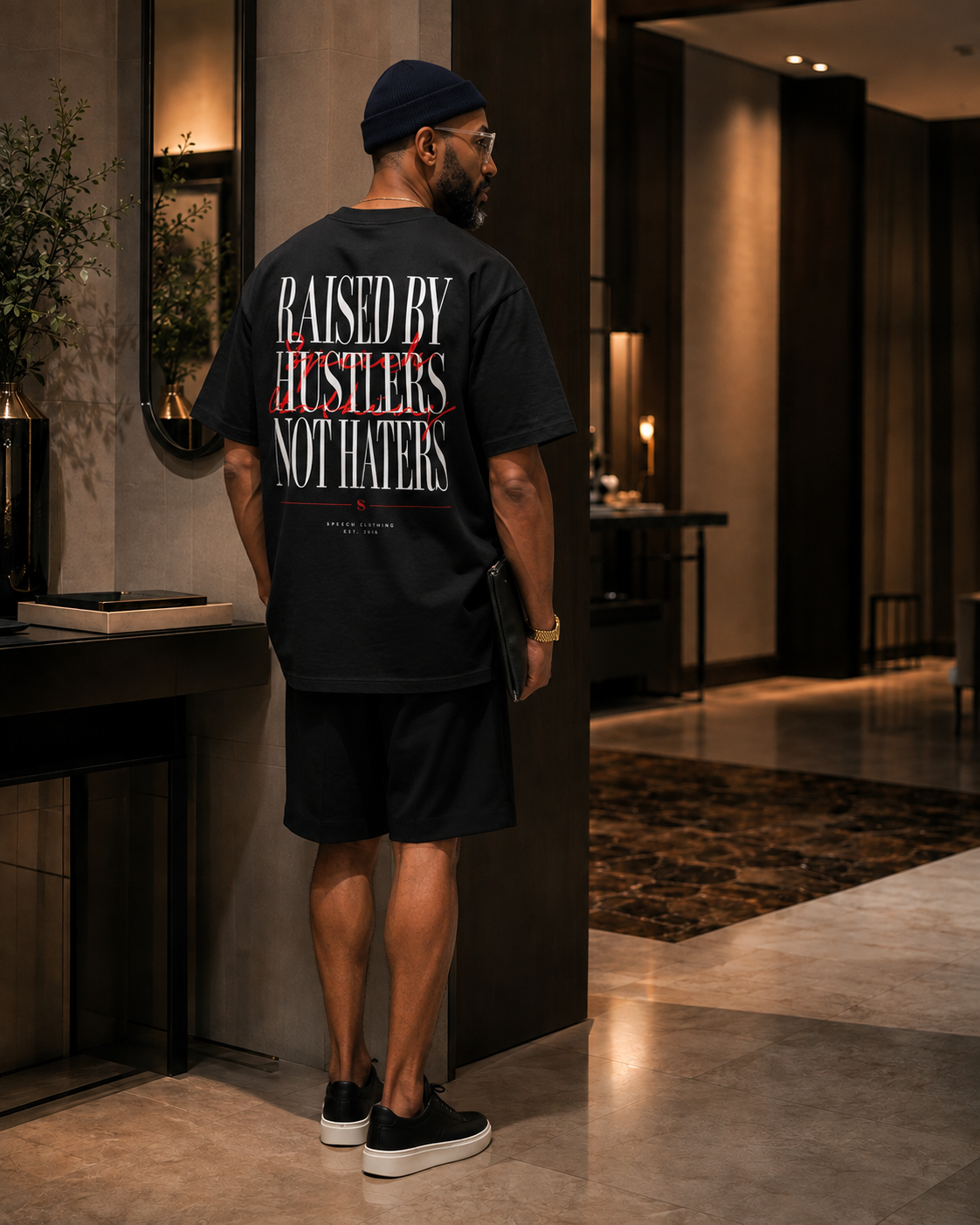 A man stands in a modern indoor hallway wearing a black graphic T-shirt from the back, which features the text 'RAISED BY HUSTLERS NOT HATERS' in white and red. He is also wearing black shorts, a black beanie, and black slip-on shoes.