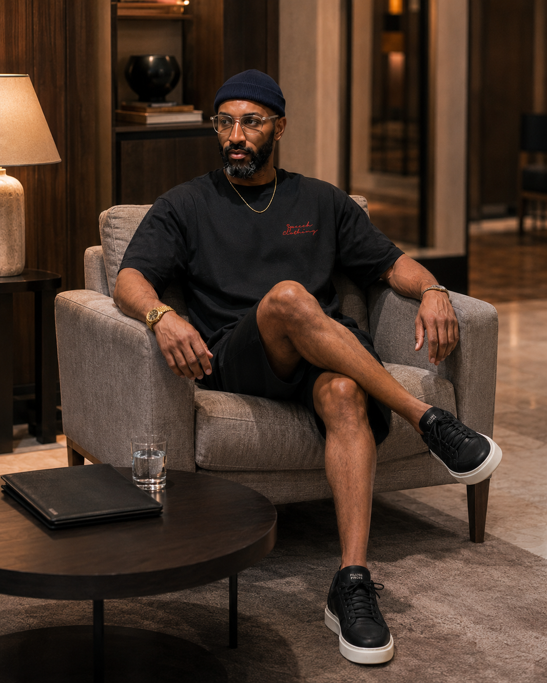 A man wearing a black t-shirt, black shorts, and black sneakers sits in a gray armchair in a modern, upscale living room setting.