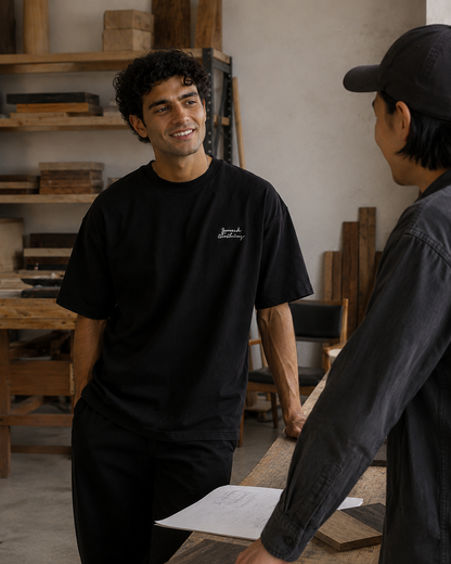 A man in a black t-shirt with a small white script logo on the left chest stands in a workshop, smiling and talking to another person wearing a black cap and dark gray shirt.