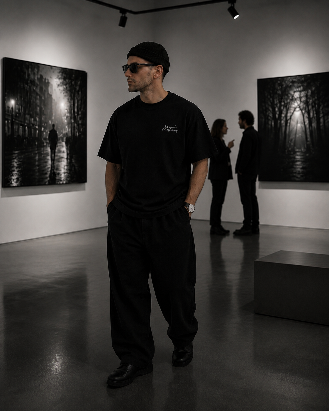 A man wearing a black t-shirt, black sweatpants, and a black beanie stands in a gallery room with black and white art prints on the walls. The t-shirt has a small white script logo on the left chest.