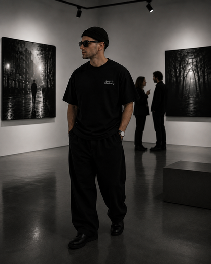 A man wearing a black t-shirt, black sweatpants, and a black beanie stands in a gallery room with black and white art prints on the walls. The t-shirt has a small white script logo on the left chest.