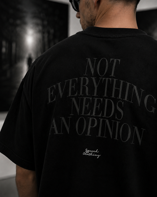 A man viewed from behind wearing a black oversized T-shirt with large gray text reading 'NOT EVERYTHING NEEDS AN OPINION' and a small white script logo near the lower back.