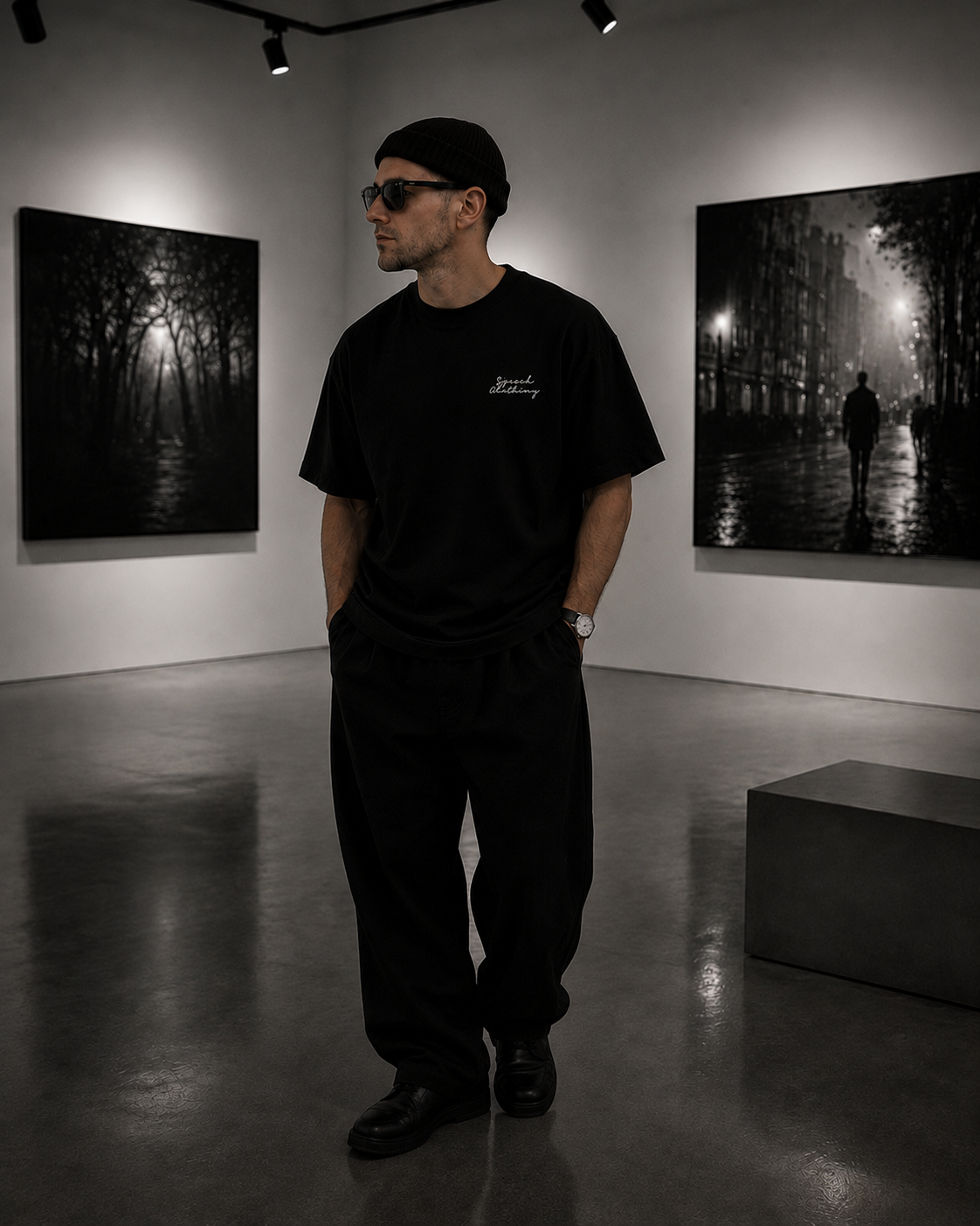 A man wearing a black t-shirt with a small white script logo on the chest, black sweatpants, and a black beanie stands in a gallery room with black and white art prints on the walls.