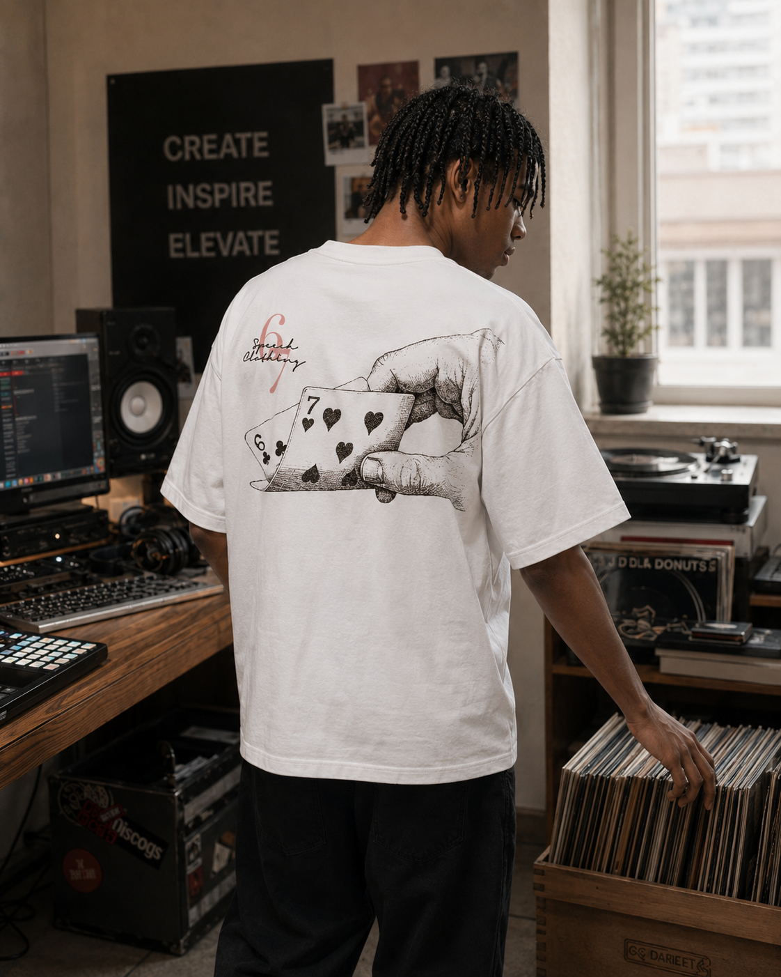 A man stands in a music studio wearing a white graphic t-shirt, showing the back of the shirt which features a black line art illustration of a hand holding playing cards. The studio is filled with recording equipment, speakers, and vinyl records.