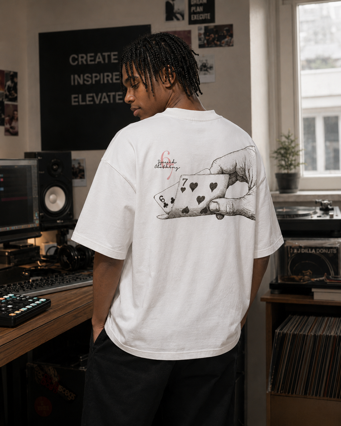 A man wearing a white graphic t-shirt with a large black illustration of a hand holding playing cards on the back, standing in a music studio room.
