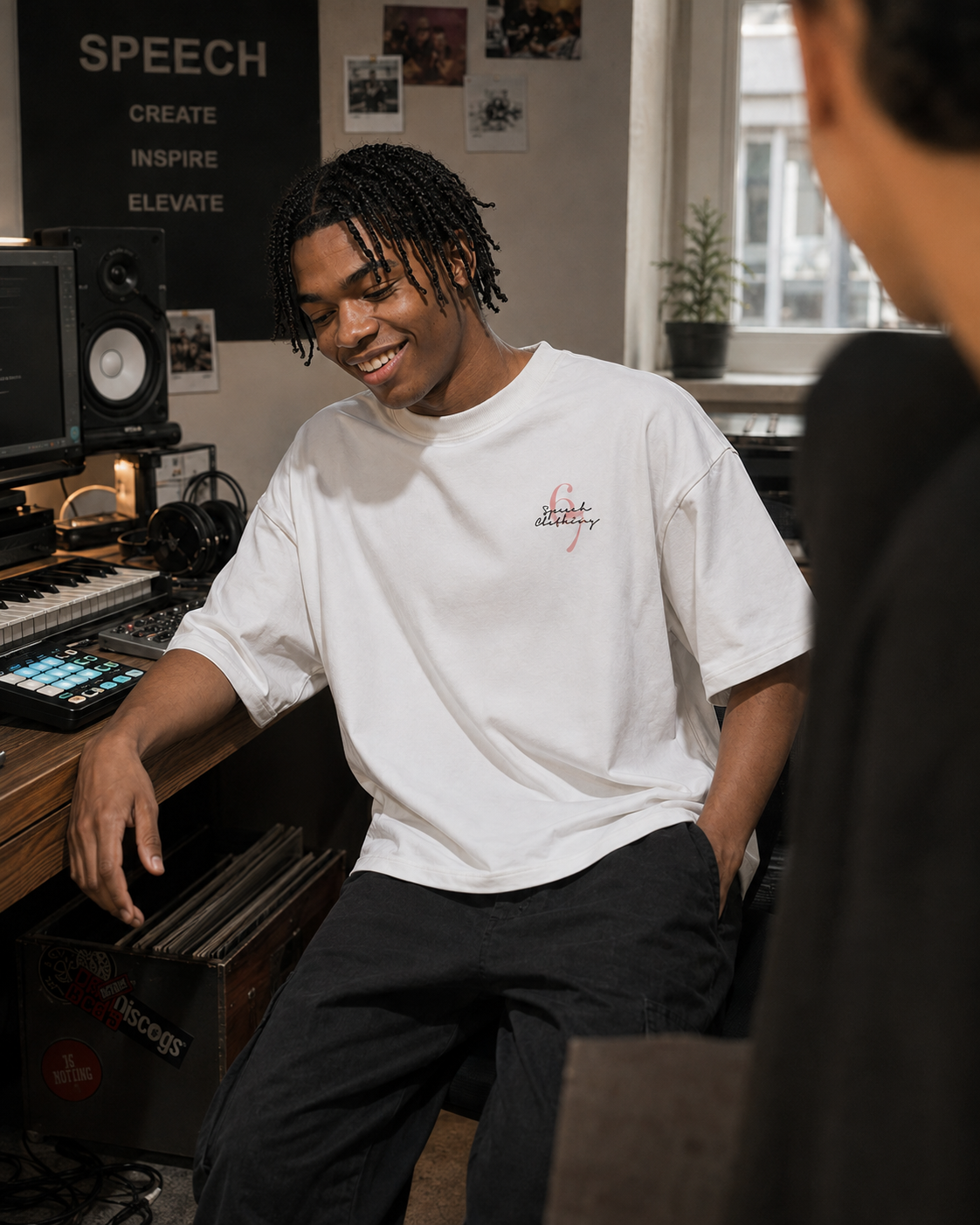 A man in a music studio wearing a white t-shirt with a small red and black graphic on the left chest, paired with dark gray sweatpants. The background features audio equipment, speakers, and a window.
