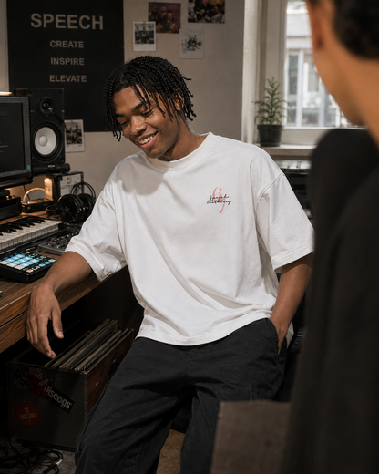 A man in a music studio wearing a white t-shirt with a small red and black graphic on the left chest, paired with dark gray sweatpants. The background features audio equipment, speakers, and a window.