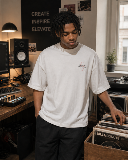 A man with black dreadlocks stands in a music studio wearing a white crewneck t-shirt with a small red and black graphic on the left chest, paired with black pants. He is reaching for vinyl records in a wooden crate next to him.