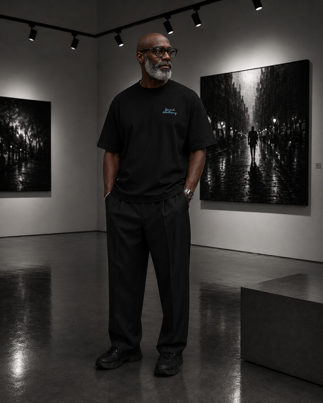A man wearing a black crewneck t-shirt with a small blue logo on the left chest and black trousers stands in a modern art gallery in front of a large black and white photograph of a street scene at night.