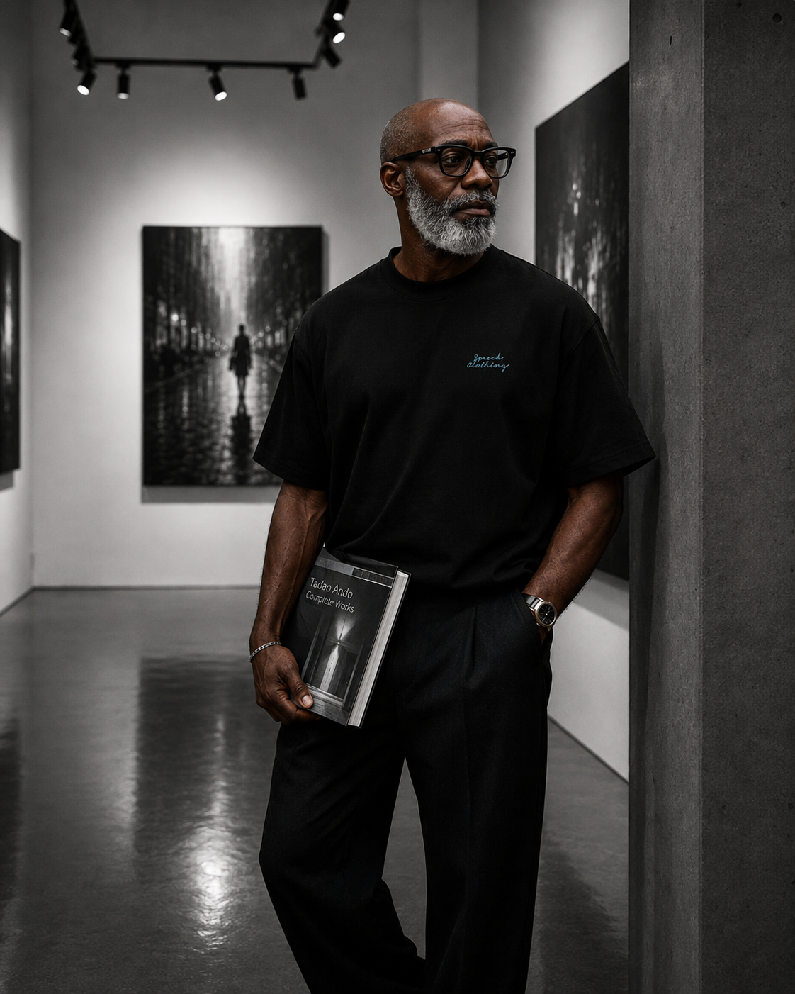 A man with a gray beard and glasses stands in a gallery reading a book, wearing a black t-shirt with a small blue script logo on the left chest and black trousers.