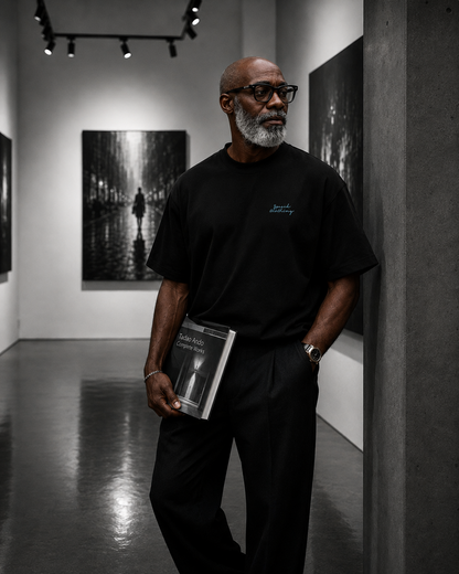 A man with a gray beard and glasses stands in a gallery reading a book, wearing a black t-shirt with a small blue script logo on the left chest and black trousers.
