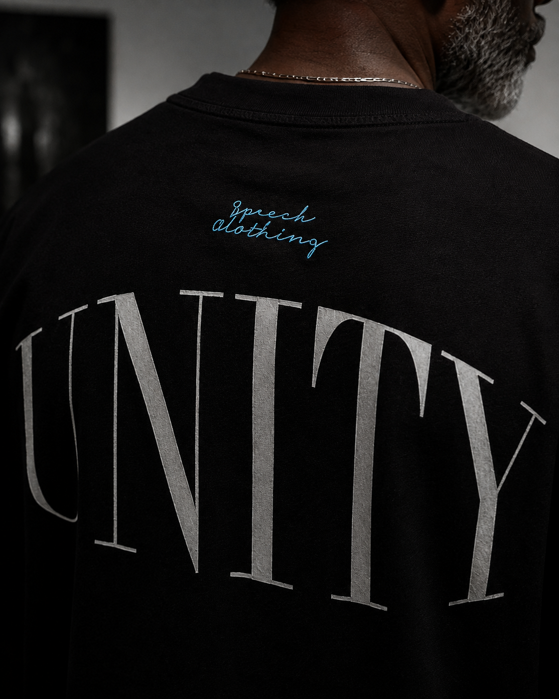 Close-up back view of a person wearing a black T-shirt with large metallic silver 'UNITY' lettering and small blue script text reading 'Speech Clothing' near the upper back.