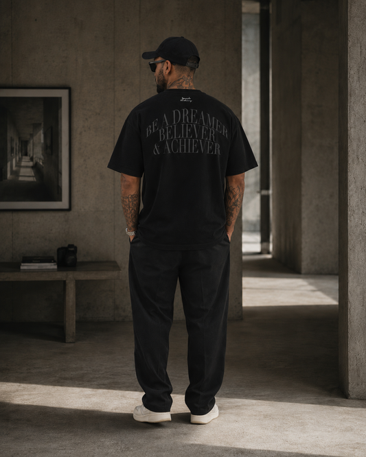 A man stands with his back to the camera wearing a black t-shirt with 'BE A DREAMER BELIEVER & ACHIEVER' printed in gray on the back, paired with black pants and white sneakers in a concrete hallway.