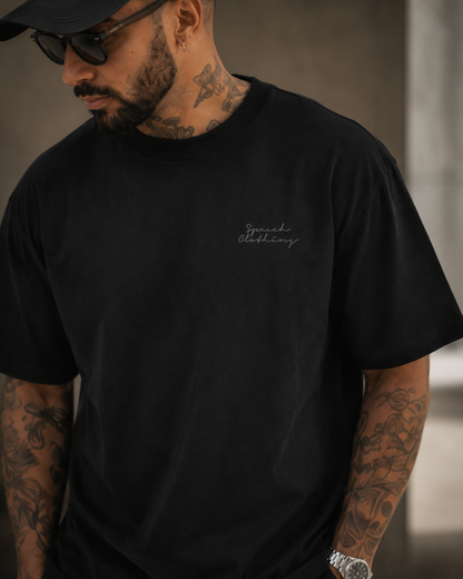 A man wearing a black crewneck t-shirt with a small white script logo on the left chest, styled with a black cap and sunglasses.