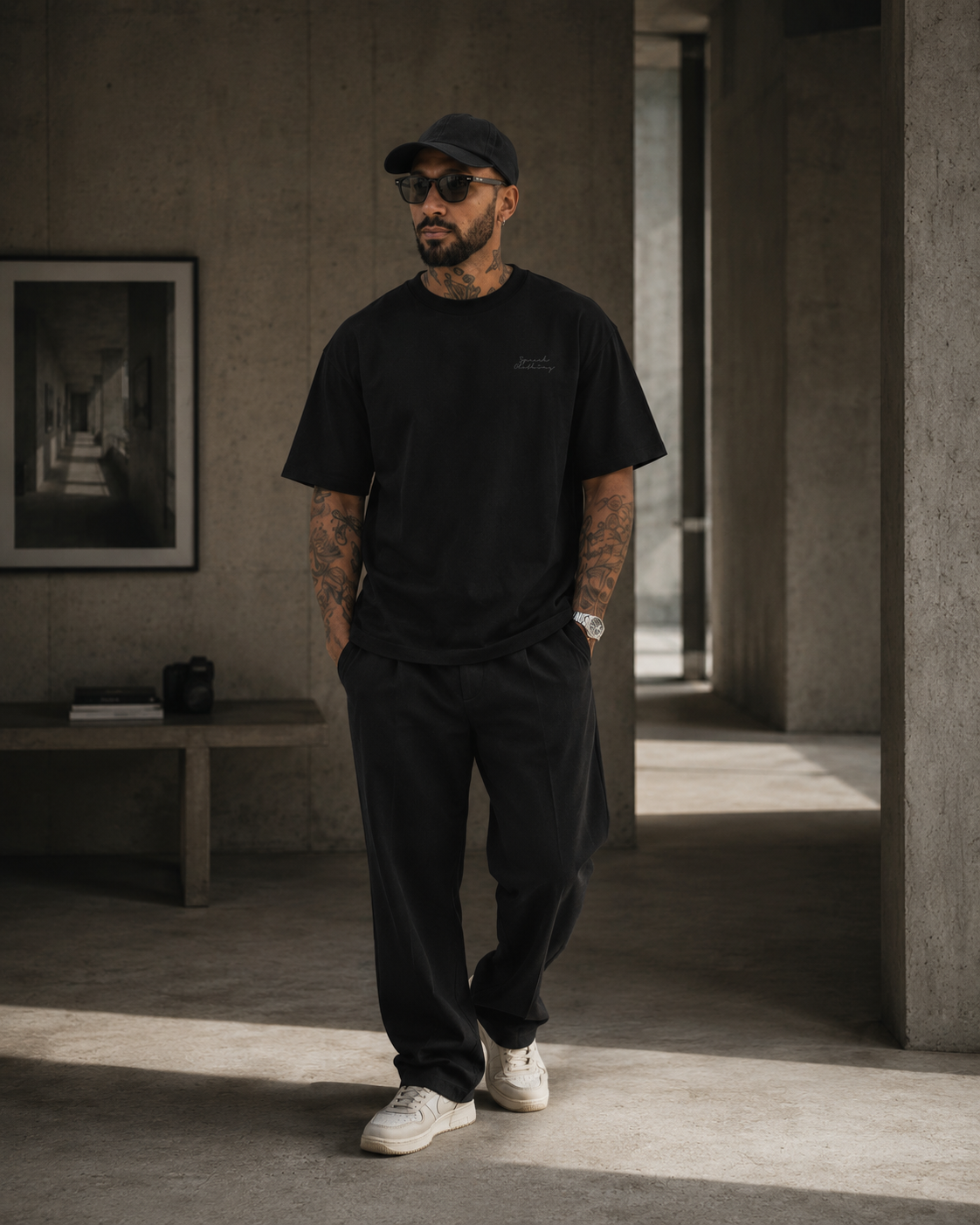 A man wearing a black crew-neck t-shirt, black sweatpants, a black baseball cap, and white sneakers, standing in a modern concrete-walled interior.