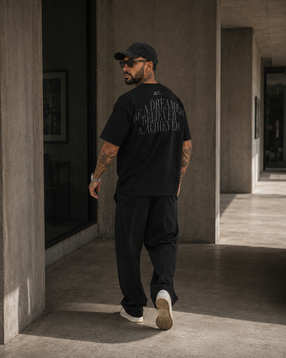 A man wearing a black t-shirt with a gray graphic text on the back, black pants, and a black cap, walking in a modern concrete corridor.
