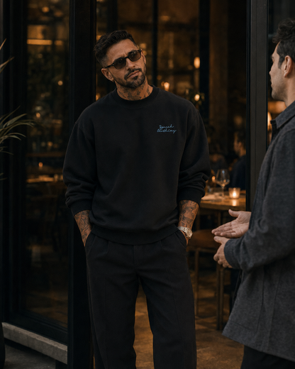 A man wearing a black crewneck sweatshirt with a small white embroidered logo on the left chest, paired with black pants, standing in a dimly lit restaurant or cafe setting.