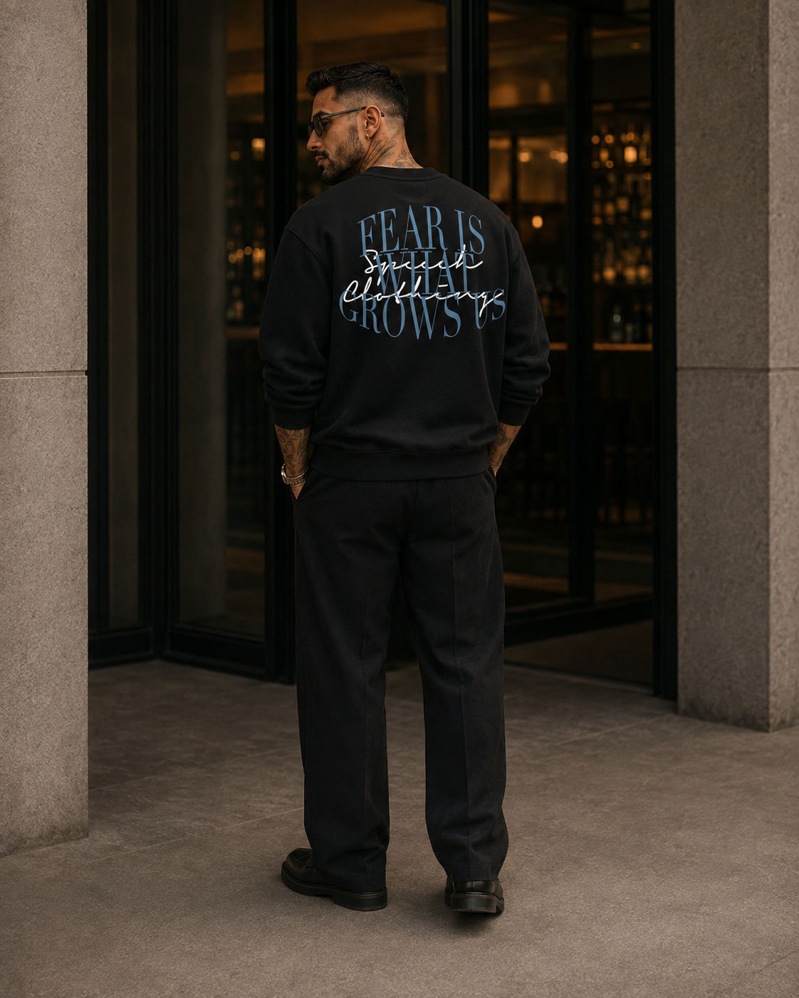 A man stands with his back to the camera wearing a black crewneck sweatshirt with a graphic on the back that reads 'FEAR IS GROWING' in blue and white script. He is wearing matching black pants and standing in front of a modern building entrance with stone columns and glass doors.