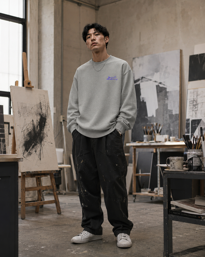 A man stands in an art studio wearing a light gray crewneck sweatshirt with a small blue graphic on the left chest, dark pants, and white sneakers. The studio features an easel with a canvas, paintbrushes, and art supplies.