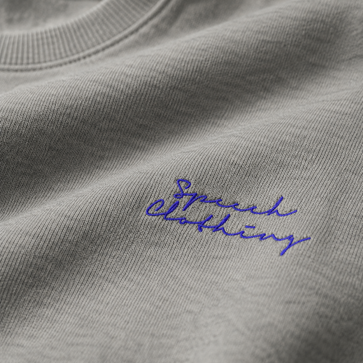 Close-up of a light gray heather crewneck sweatshirt with blue embroidered script reading 'Speech Clothing' on the chest.