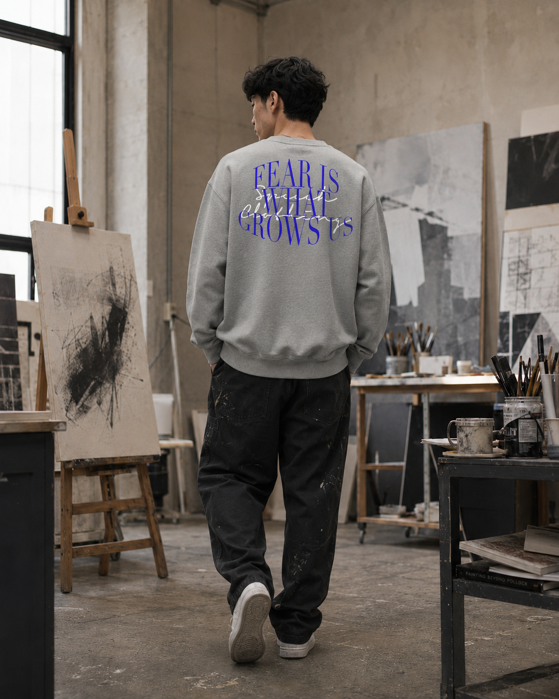 A man stands with his back to the camera in an artist studio, wearing a light gray sweatshirt with purple text reading 'FEAR IS EVERYWHERE' and dark pants.