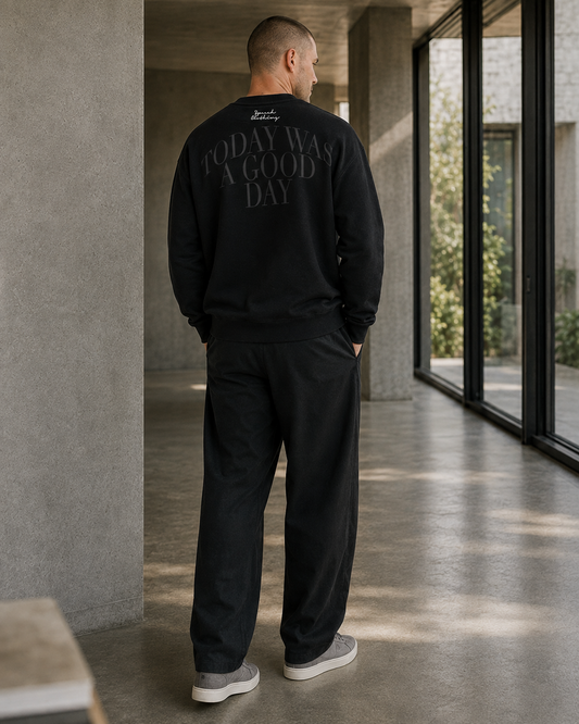 A man stands with his back to the camera in a modern concrete corridor, wearing a black crewneck sweatshirt with 'TODAY WAS A GOOD DAY' printed in gray across the back, black sweatpants, and gray slip-on shoes.