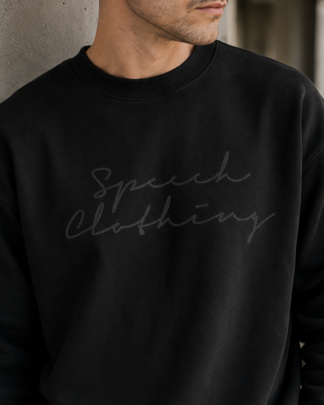 A close-up of a man wearing a black crewneck sweatshirt with 'Speech Clothing' printed in a dark gray cursive font across the chest.