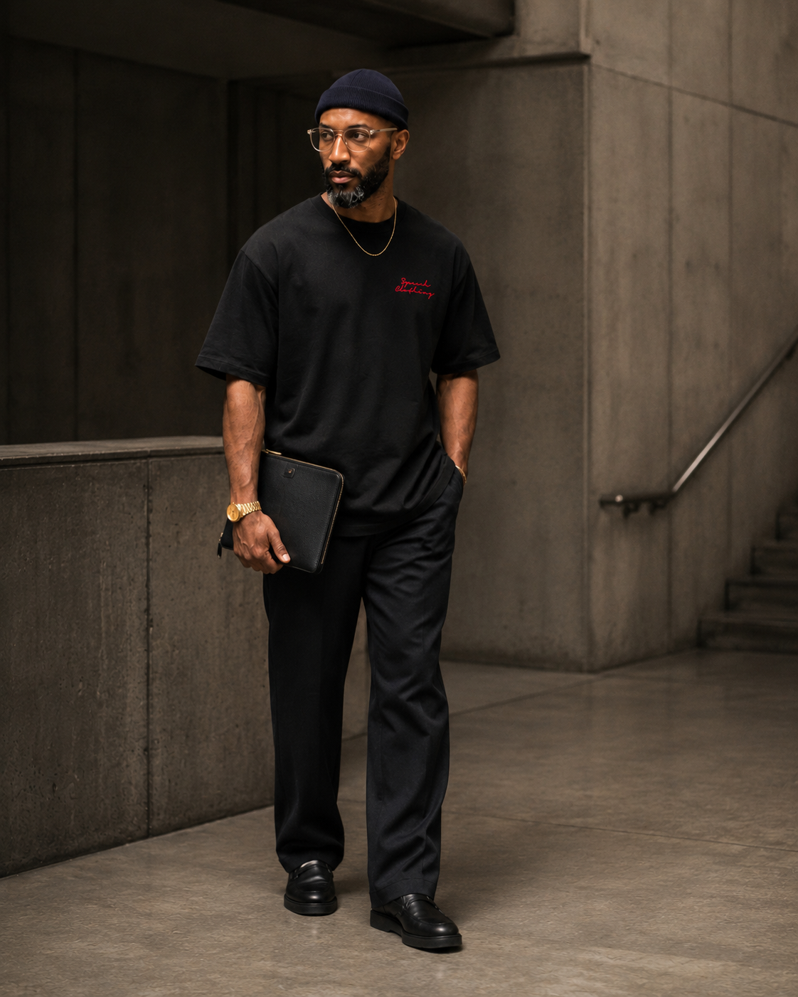 A man wearing a black crew-neck t-shirt with a small red graphic on the left chest, black trousers, and a black beanie, standing in a concrete hallway and holding a black tablet.
