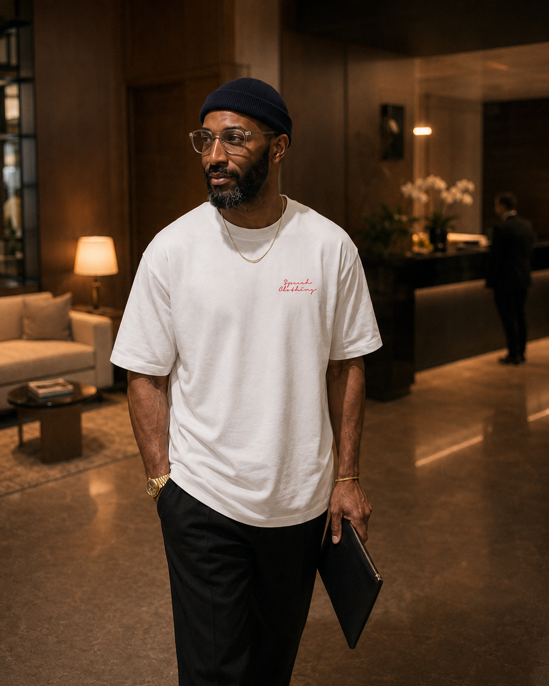A man wearing a white crewneck t-shirt with a small red embroidered logo on the chest, paired with black trousers and a dark beanie, standing in a modern, warmly lit lobby.