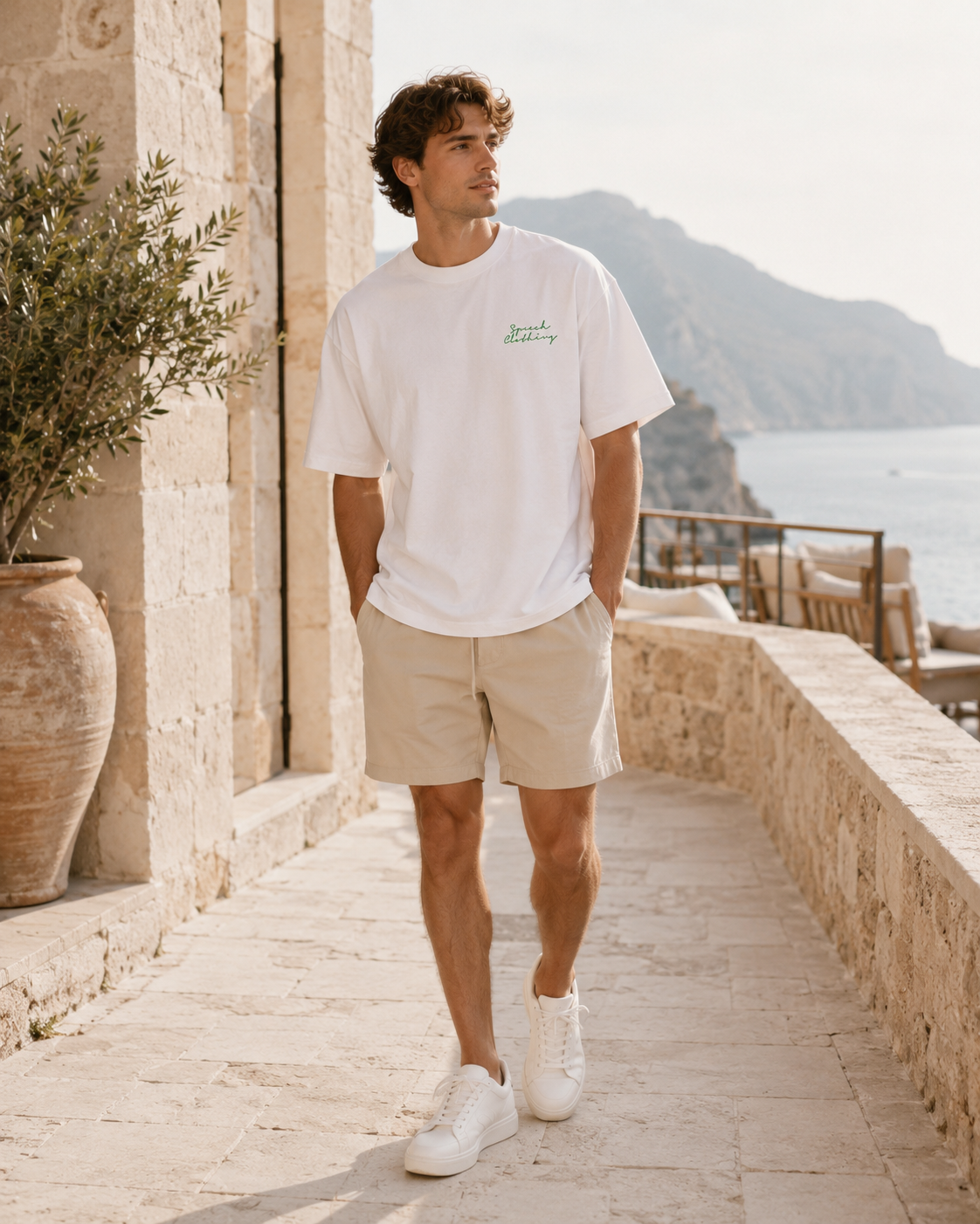 A man wearing a white crewneck t-shirt with green script embroidery, beige shorts, and white sneakers walks along a stone path with a scenic Mediterranean coastline in the background.