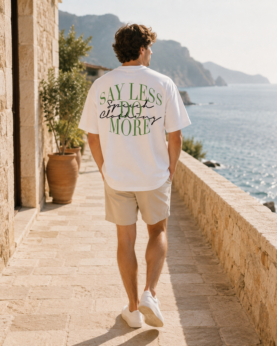 A man wearing a white graphic t-shirt and beige shorts walks along a sunny stone path by the sea, viewed from behind. The back of the t-shirt features green and black text that reads 'SAY LESS, SMILE MORE'.