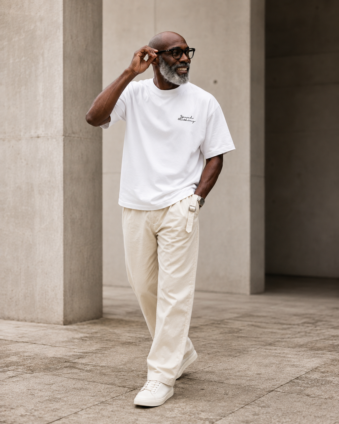 A man wearing a white crew-neck t-shirt with a small black script logo on the left chest, paired with cream-colored trousers and white sneakers, walking in a modern concrete architectural setting.