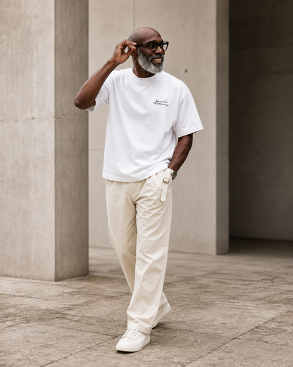 A man wearing a white crew-neck t-shirt with a small black script logo on the left chest, paired with cream-colored trousers and white sneakers, walking in a modern concrete architectural setting.
