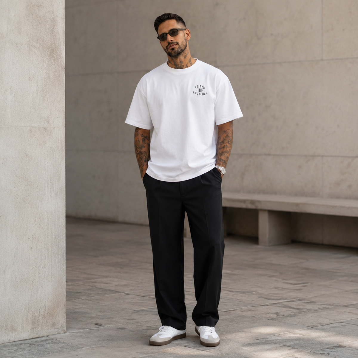 A man wearing a white crew-neck t-shirt with a small black graphic on the left chest, paired with black trousers and white sneakers, standing in a modern concrete corridor.